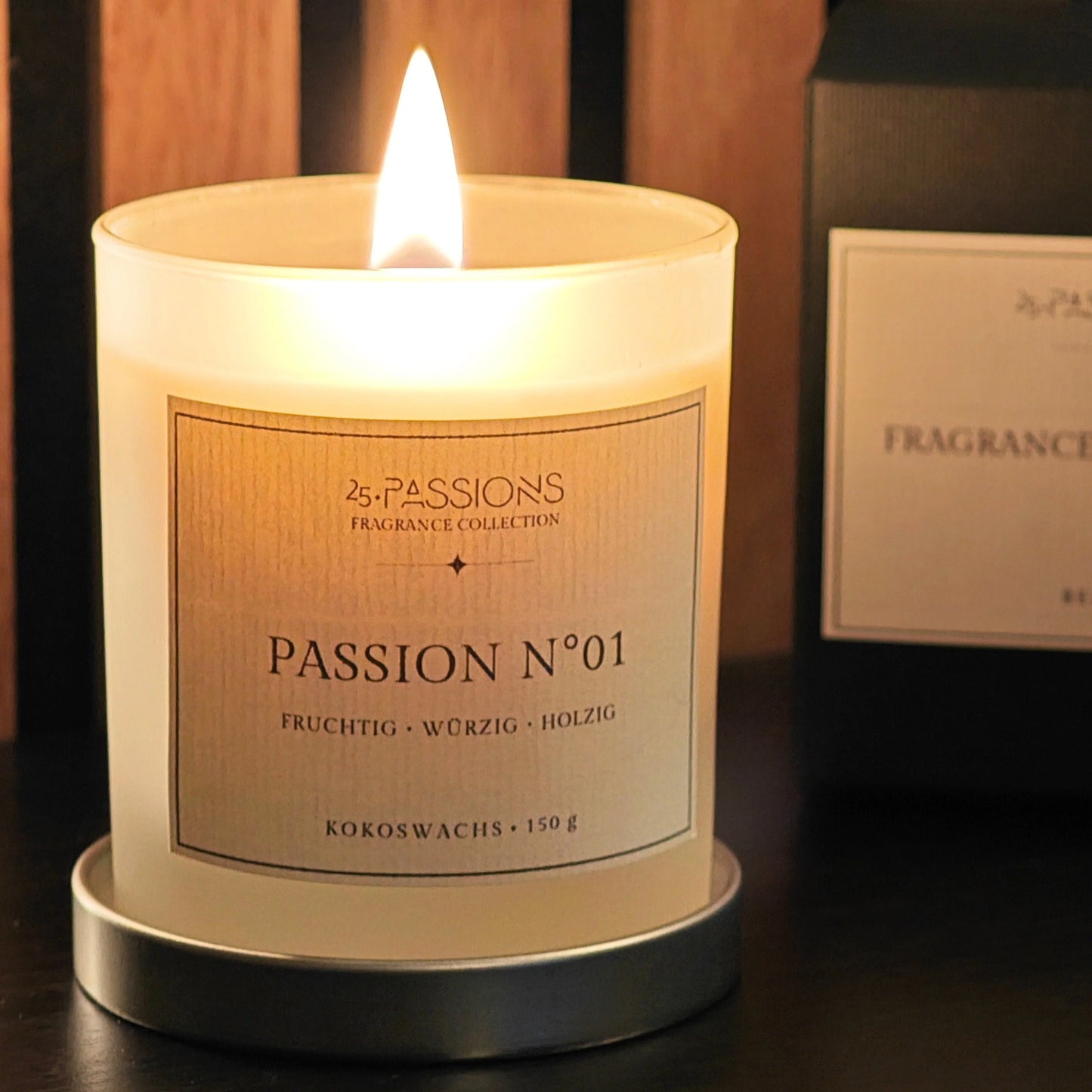 Brennende Kerze Passion N°01 von 25·PASSIONS im satinierten Glas, stehend auf dem Metalldeckel, mit Etikettangabe zu Wachsart und Füllmenge, im Hintergrund die passende Produktverpackung der Fragrance Collection